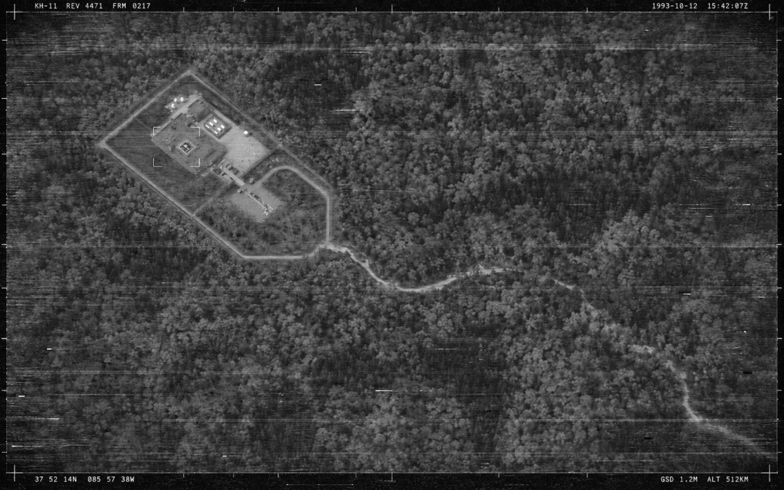 A grainy 1990s satellite frame of a fenced compound in dense autumn forest: a flat-roofed building with a helipad on the roof, a car park, a loop road inside the fence and a long dirt track winding away to the south-east; frame readouts, a timestamp of 12 October 1993 and coordinates in the corners.
