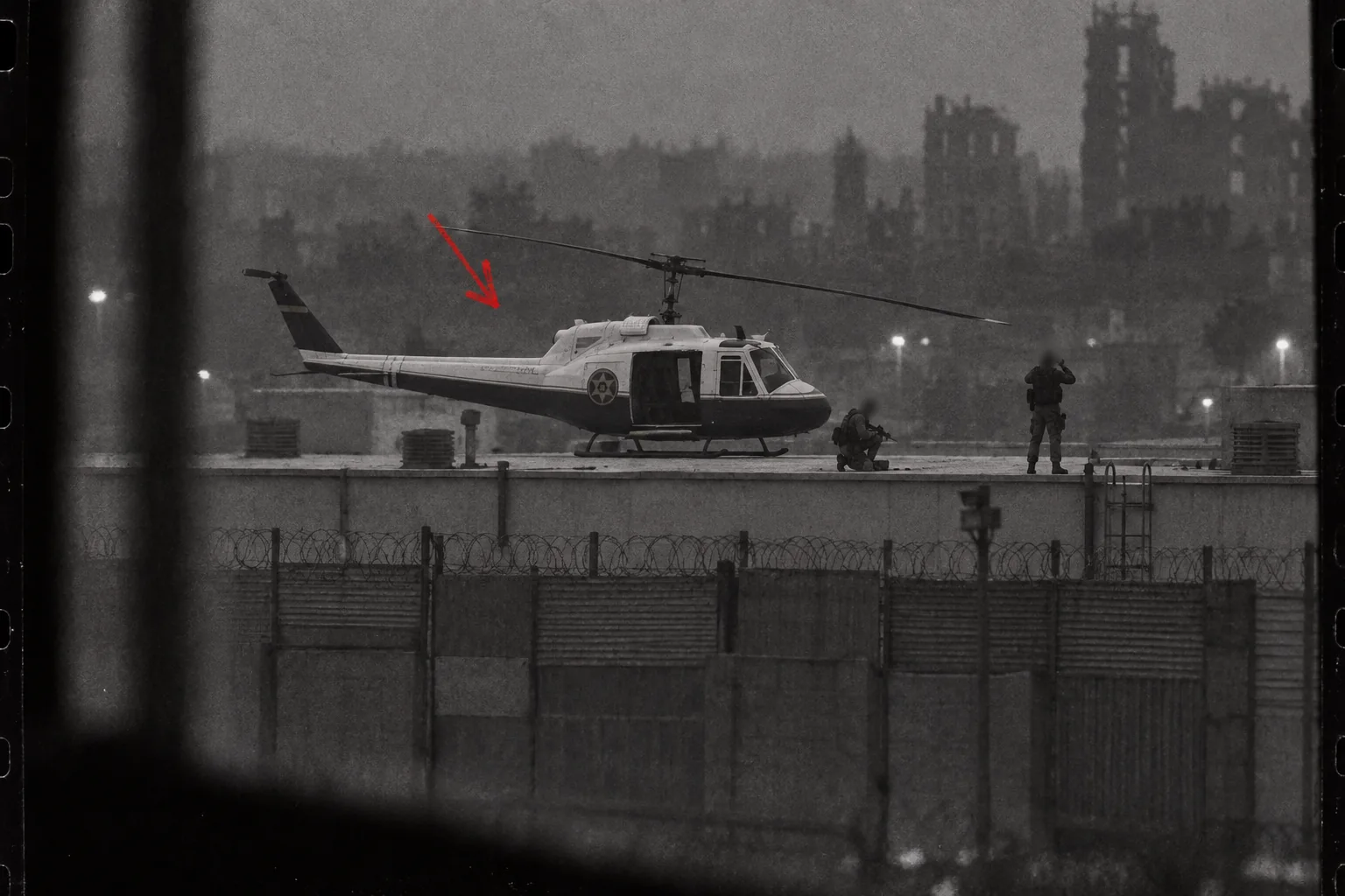 A grainy black-and-white long-lens photograph taken from behind a window frame at dusk: a police Huey in a pale-over-dark paint scheme with a star badge sits on the flat roof of a warehouse behind a towering wall of corrugated steel and razor wire, one armed figure kneeling by it and another standing with binoculars, both faces blurred out, a red grease-pencil arrow drawn on the print.