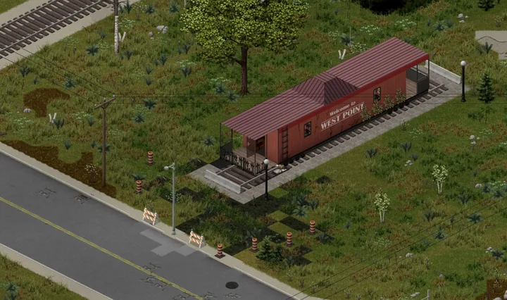 A game screenshot from above of a red caboose on a short length of track beside a road, Welcome to West Point painted on its side, patches of burned black grass around it and orange roadwork barrels and striped barriers on the verge.