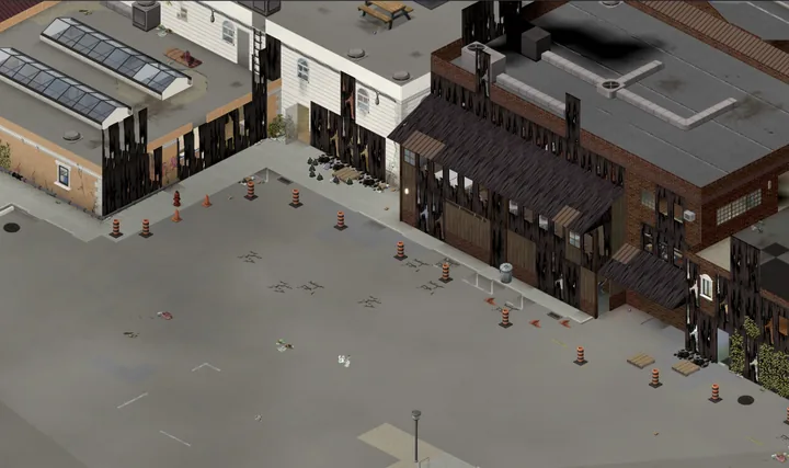A game screenshot from above of a big empty car park behind a row of shops: a flat roof with skylights, a white wall with arched windows and a brick building with a timber porch front, all streaked black by fire, orange roadwork barrels dotted across the tarmac.